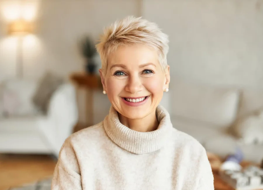 Smiling mature woman with short blonde hair wearing a beige turtleneck sweater, showcasing healthy teeth after Dental Fillings at Gridley Dental